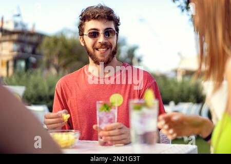 A young man is sitting at an outdoor pub table, smiling and having a great time with his friends (off-camera). He is dressed in a red t-shirt and hold Stock Photo