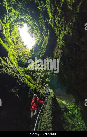 Tourists descend into the Algar do Carvão, a vertical lava tube dropping 300 feet from the surface accessible by a narrow staircase to a clear water pool at the bottom in the central mountains, Terceira Island, Azores, Portugal. Stock Photo