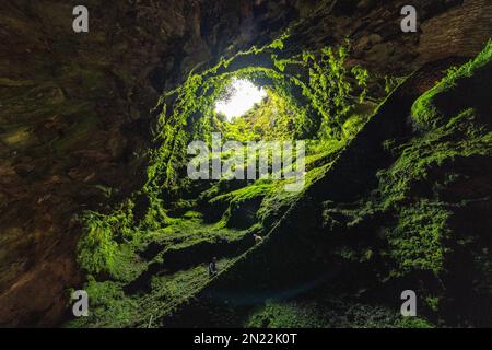 Tourists descend into the Algar do Carvão, a vertical lava tube dropping 300 feet from the surface accessible by a narrow staircase to a clear water pool at the bottom in the central mountains, Terceira Island, Azores, Portugal. Stock Photo