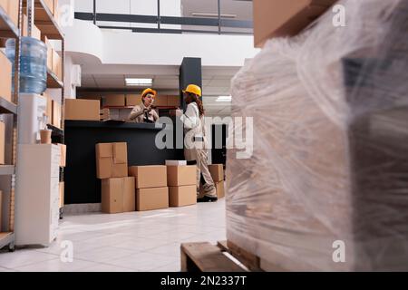 Retail storehouse operative managing product receiving and scanning suticase at desk. Two diverse employees wearing protective overalls working in warehouse full of cardboard boxes parcels Stock Photo