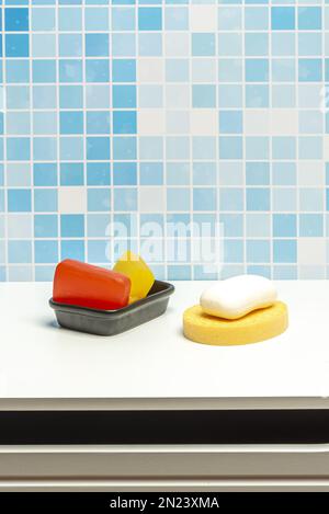 Black porcelain bowl with glycerin soap bars, a white soap bar and a yellow sponge on a white surface for hand washing in a blue tiled bathroom Stock Photo