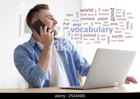 Young man talking on mobile phone while working with laptop at desk, word cloud in front of him Stock Photo