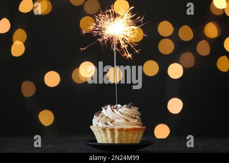 Cupcake with burning sparkler on black table against blurred festive lights Stock Photo