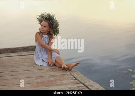 Cute little girl wearing wreath made of beautiful flowers on pier near river Stock Photo