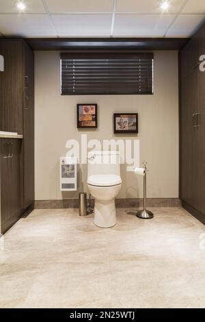 White porcelain toilet and brown wood veneer cabinets in laundry room bathroom in basement inside small home. Stock Photo