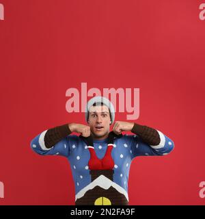 Surprised man in hat pulling decorative Santa on his Christmas sweater against red background Stock Photo