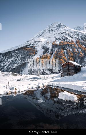 Swiss chalet in a wintry landscape with golden larches and snowy mountains reflecting in a small mountain lake in Arolla, Wallis, Switzerland Stock Photo