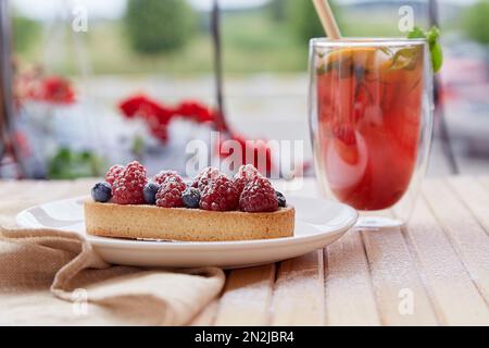 Refreshing raspberry- orange lemonade with mint and cane straw outside. Sugar free tart with raspberry and blueberry at the terrace on the wooden tabl Stock Photo