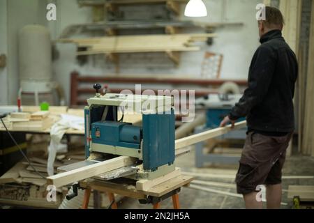 Guy is chipping board on machine. Worker makes furniture. Work in carpentry workshop. Stock Photo