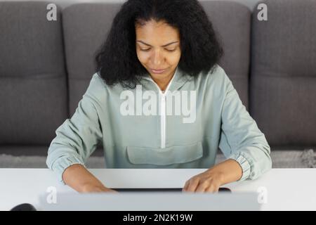 Young adult POC woman works on computer at home. Overhead photo of focused black female person doing distant work on laptop Stock Photo