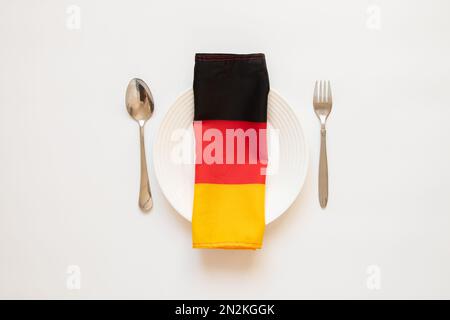 The flag of Germany lies on a white plate next to a spoon and fork on a white background, the national flag of Germany Stock Photo