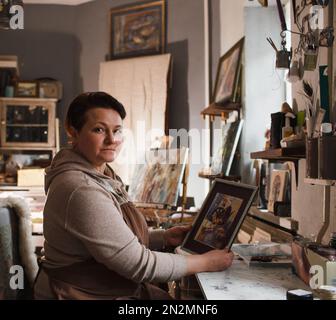 the artist girl in the studio holds a framed painting in her hands. Stock Photo