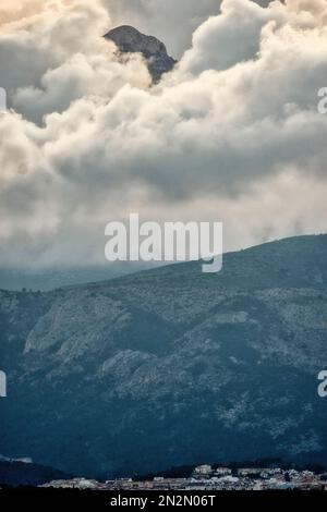 mountaintop peeking through fluffy cumulus clouds above village in Spain Stock Photo