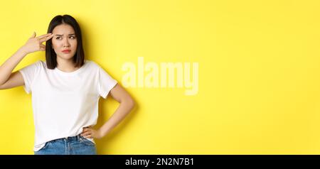 Woman showing kill me please gesture, shooting herself in head with finger gun from boredom, standing upset over yellow background Stock Photo