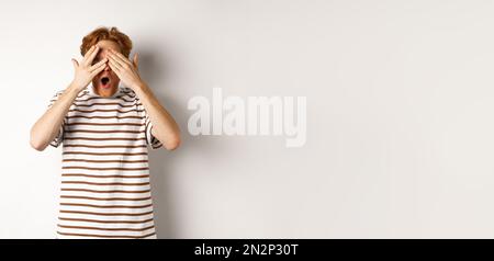 Shocked and embarrassed redhead guy covering eyes, peeking through fingers and gasping, standing over white background Stock Photo