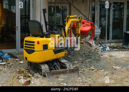 Small yellow excavator and concrete mixer at a construction site in front of a building under construction. Stock Photo