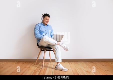 Happy asian man in headphones having video call on laptop, sitting on chair against white wall, full length, free space Stock Photo