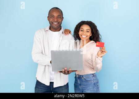 Happy black middle aged man and young woman showing credit card and laptop, standing over blue studio background Stock Photo