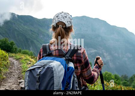 rear view of young woman in plaid shirt and cap with big backpack with trekking poles walking along mountain trail in summer hiking healthy active lif Stock Photo