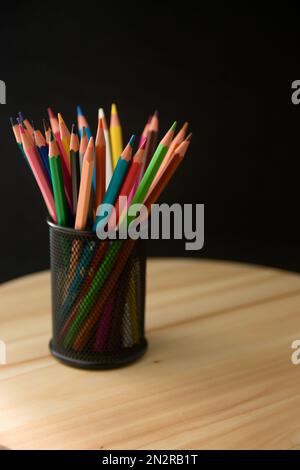 Closeup of a black metallic pot with colored pencils of various colors on a wooden table and black background with copy space Stock Photo