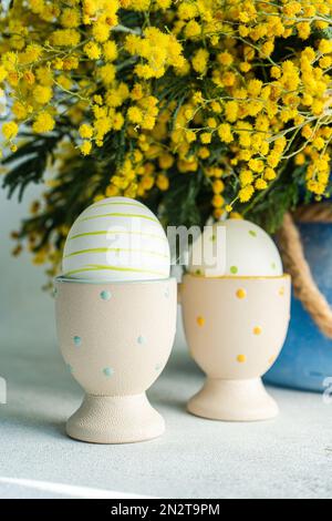 Painted Easter eggs in egg cups next to a bunch of yellow mimosa flowers Stock Photo