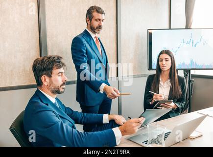 Group of businesspeople in formal clothes gathering at table with laptop and working with charts during meeting in office Stock Photo