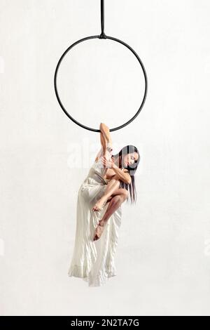 Full body graceful barefoot female gymnast in dress with dark hair crossing legs and performing elbow grip pose on aerial ring against white backgroun Stock Photo