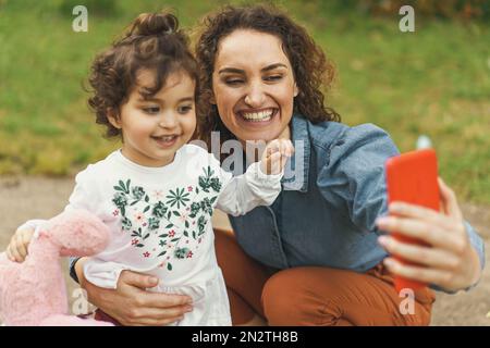 A young mother and her two-year-old daughter play and take a selfie with their smartphone in the park - family people lifestyle concept Stock Photo