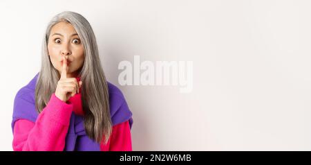 Close-up of stylish senior asian woman in hipster outfit telling hush, shushing at looking at camera, show taboo gesture, standing over white Stock Photo