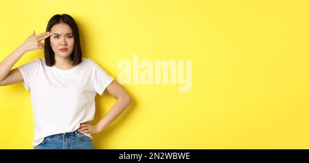 Woman showing kill me please gesture, shooting herself in head with finger gun from boredom, standing upset over yellow background Stock Photo