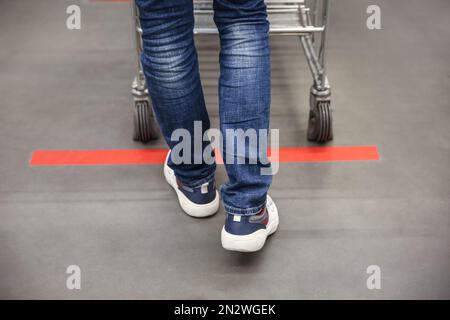 Person with shopping cart standing behind taped floor marking in store for social distance, closeup. Preventive measure during coronavirus pandemic Stock Photo
