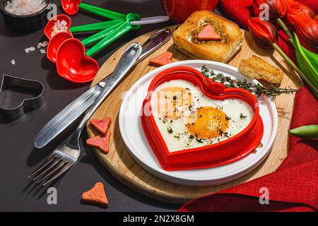 Heart-shaped fried egg served with toasted bread and spices on a plate. Romantic art food idea for Valentine's breakfast. Hard light, dark shadow, bla Stock Photo