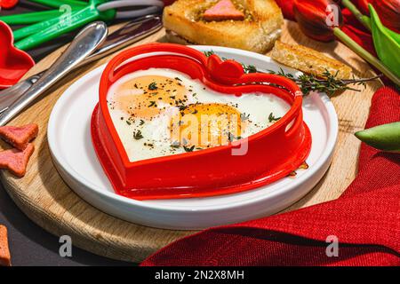 Heart-shaped fried egg served with toasted bread and spices on a plate. Romantic art food idea for Valentine's breakfast. Hard light, dark shadow, bla Stock Photo