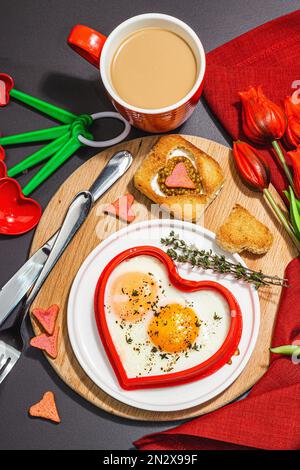 Heart-shaped fried egg served with toasted bread and spices on a plate. Romantic art food idea for Valentine's breakfast. Hard light, dark shadow, bla Stock Photo