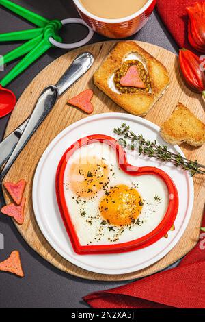 Heart-shaped fried egg served with toasted bread and spices on a plate. Romantic art food idea for Valentine's breakfast. Hard light, dark shadow, bla Stock Photo
