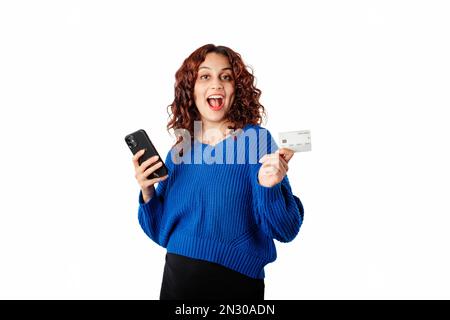 Happy woman standing isolated over white background holding a credit card and a phone looks overjoyed and looks at the camera. Seeing an incredible ca Stock Photo
