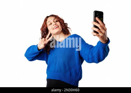 Young girl smiling happy wearing pullover sweater isolated over white background makes a video call or selfie with smartphone makes a peace symbol wit Stock Photo