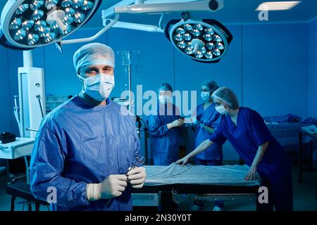 Male surgical doctor in blue scrub suit with surgical scissors in hands standing inside operating room with surgery team Stock Photo