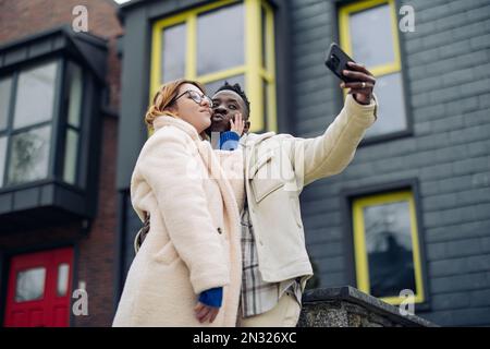 Young interracial couple takes a selfie together on street against background of houses. Concept of love relationships and unity between different hum Stock Photo