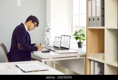 Financial accountant writing something while sitting at office desk with two laptops Stock Photo