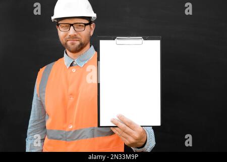 Happy Engineer Pointing At Blank Paper Sheet, Clipboard. Man Builder In Safety Vest And Hard Hat Advertising Empty White Tablet Standing Over Black Ba Stock Photo