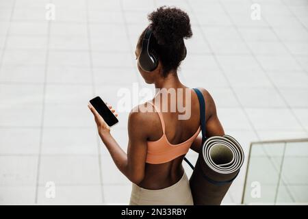 Young fit sporty afro woman with yoga mat holding phone while standing with her back Stock Photo