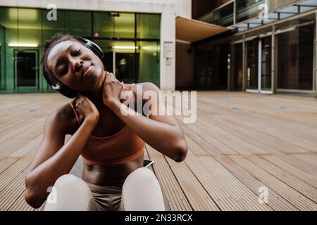 Young beautiful african woman in headphones stretching her neck with closed eyes, while sitting on mat near moderns building outdoors Stock Photo