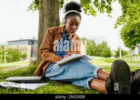 Young beautiful thoughtful african woman in headphones doing homework and touching chin while sitting in park Stock Photo