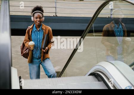 Young beautiful african woman in headphones with coffee and folder looking aside while standing near escalator outdoors Stock Photo