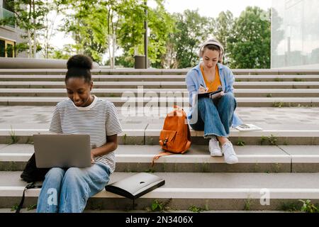 Young multiracial student women doing homework while sitting on stairs outdoors Stock Photo
