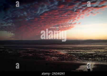 Two people walking along the beach at Cubbins Green, West Kirby, Wirral, looking over to the coast of North Wales on a February evening at sunset. Stock Photo