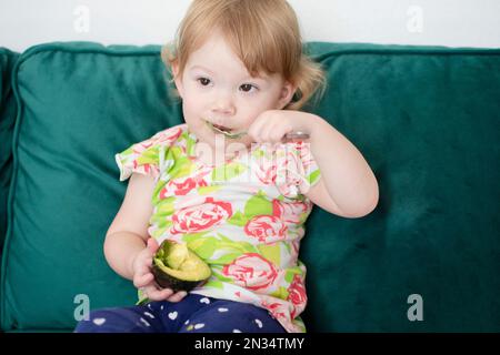 Caucasian toddler eating avocado with the spoon. Child feeding herself with a spoon. Healthy eating habits. Organic avocado. Stock Photo