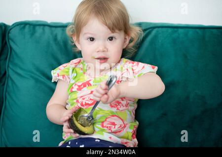 Caucasian toddler eating avocado with the spoon. Child feeding herself with a spoon. Healthy eating habits. Organic avocado. Stock Photo