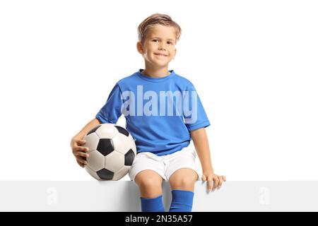 Boy in a blue soccer jersey sitting on a blank panel and holding a ball isolated on white background Stock Photo